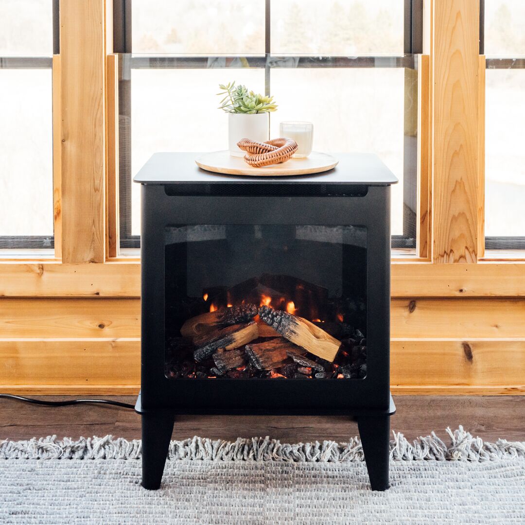 Electric stove with glowing embers and realistic flame effect, placed in front of cabin-style windows with wooden frames and a gray rug on the floor