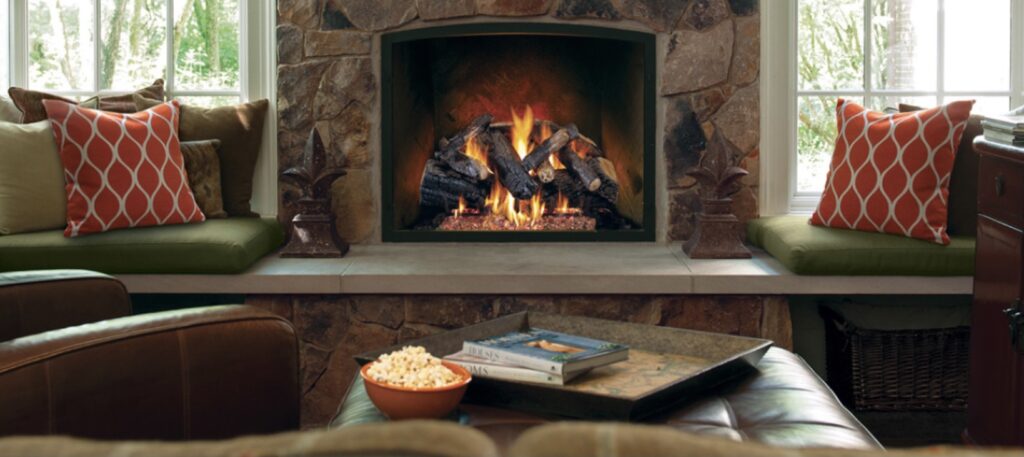Warm living room with a stone fireplace, lit fire, and built-in window seating with green cushions and orange patterned pillows.