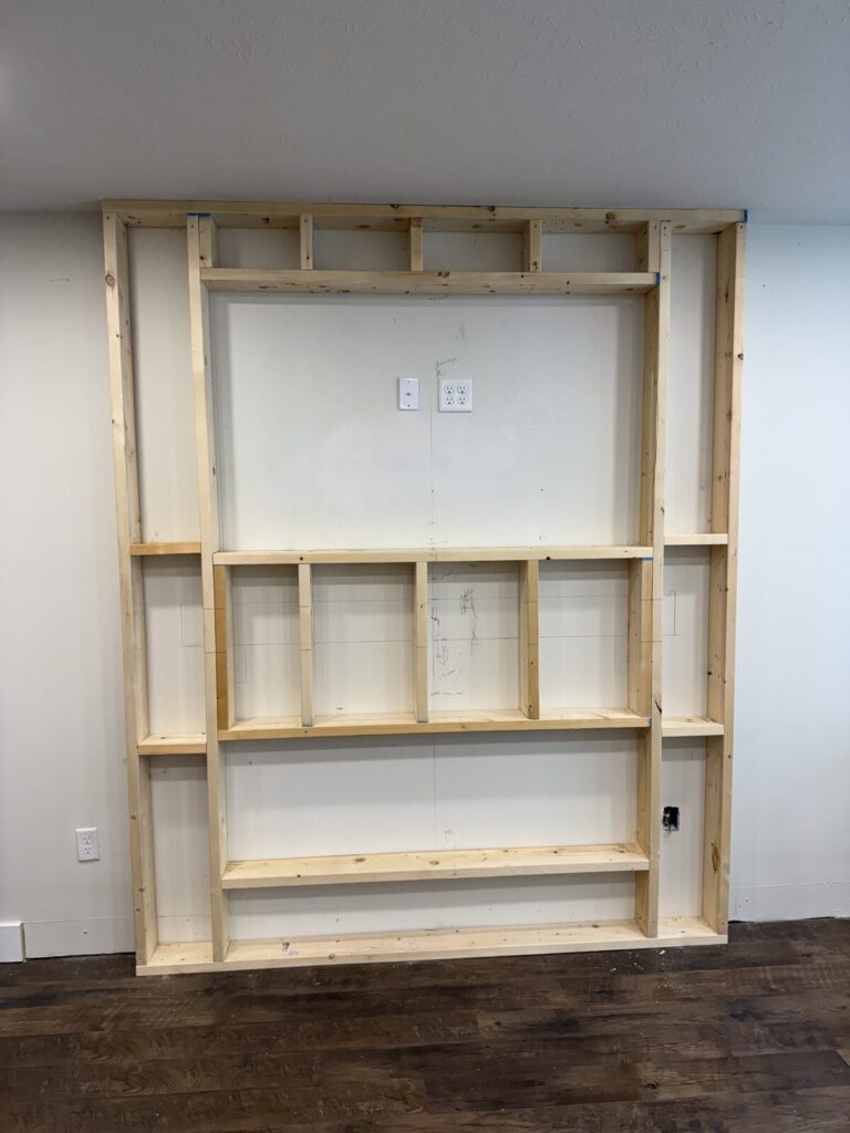 Freshly built wood stud frame on a living room wall with a recessed opening for a TV and shelves below