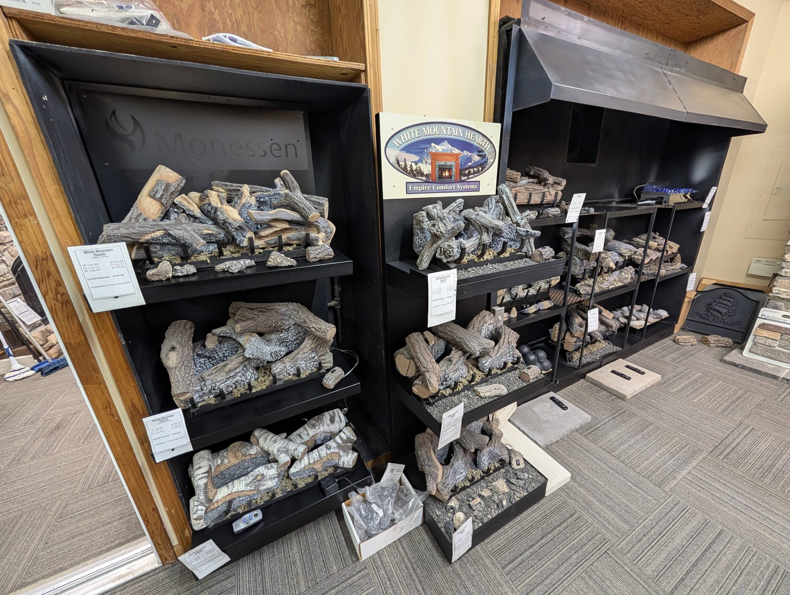 Showroom display of Monessen and White Mountain Hearth gas log sets arranged on black shelving units, featuring various realistic wood styles and ember beds