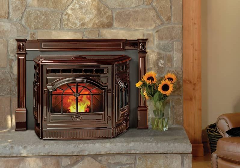 Mahogany-finished Harman pellet fireplace insert with glowing flame, set in a rustic stone surround next to a leather armchair and vase of sunflowers