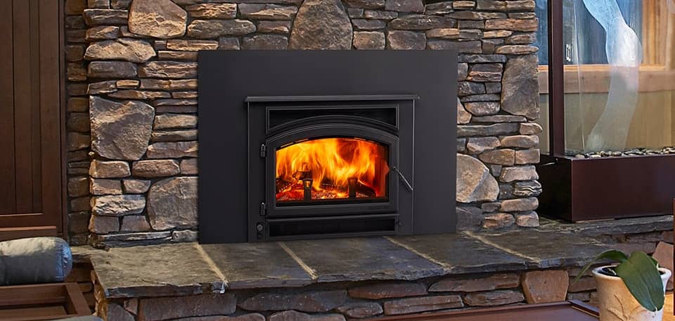 Quadra-Fire wood fireplace insert with roaring flames, installed in a rugged stone surround with a wide black steel faceplate and natural stone hearth