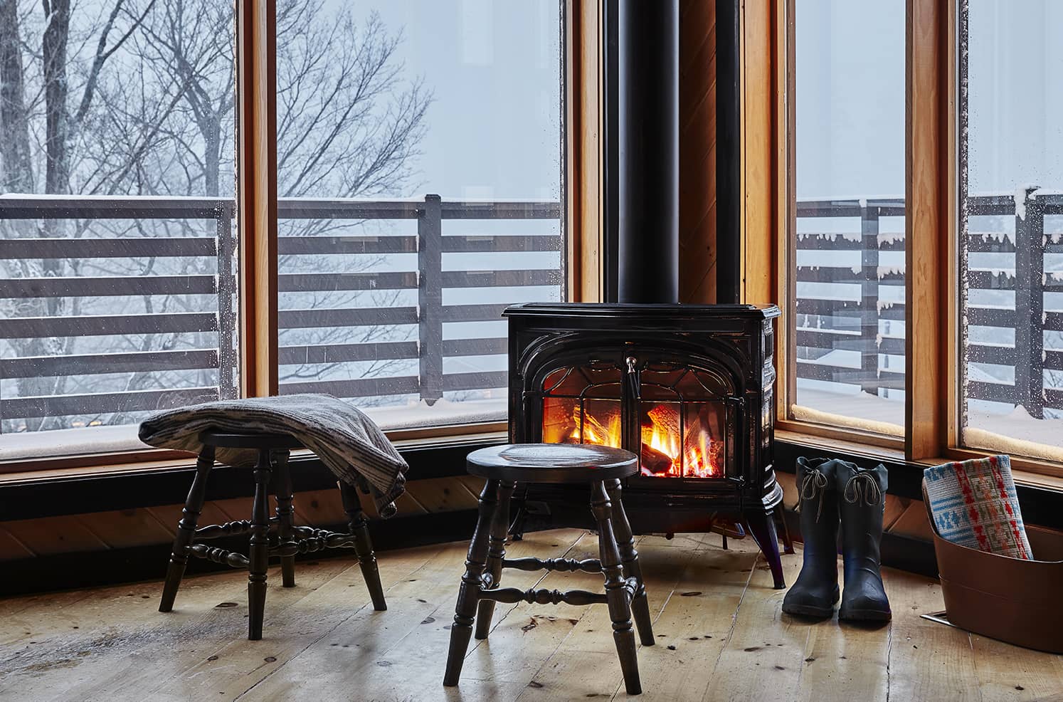 Black gas stove with glowing flames and embers in a wooden cabin living space, surrounded by stools, boots, and winter decor near large snowy windows