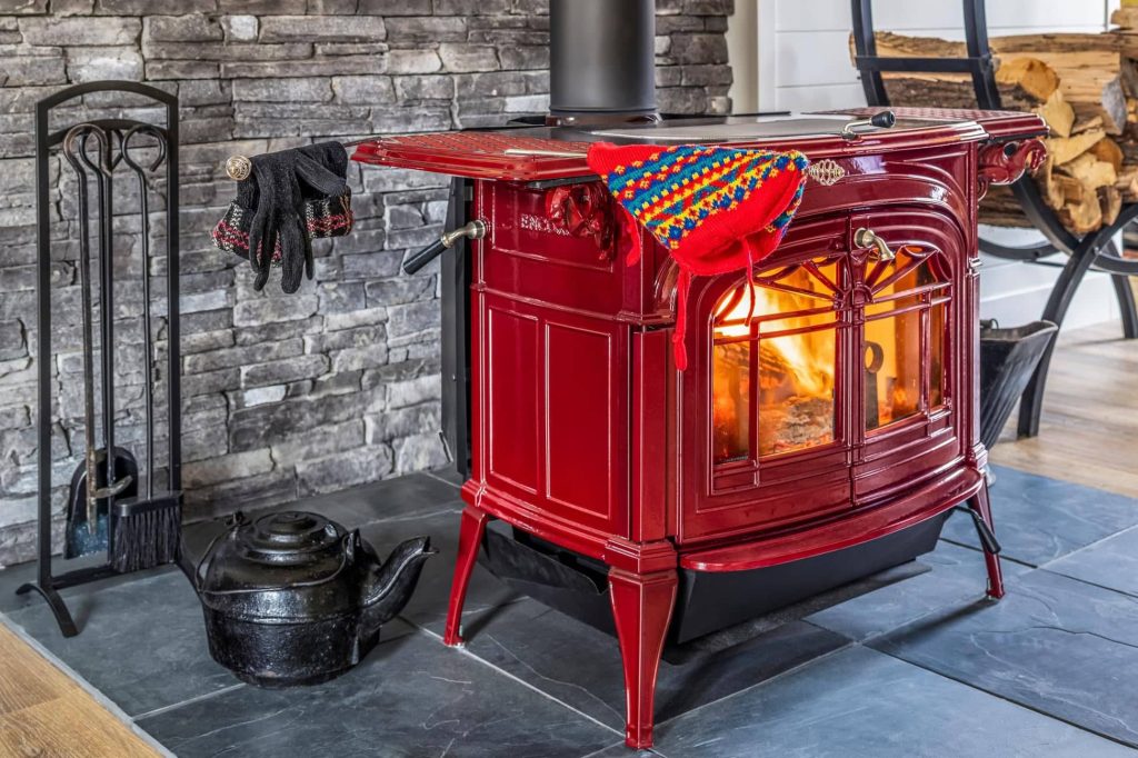 Glossy red Vermont Castings wood stove burning brightly on a slate hearth, with fire tools, kettle, gloves, and a colorful knit hat against a stacked stone wall